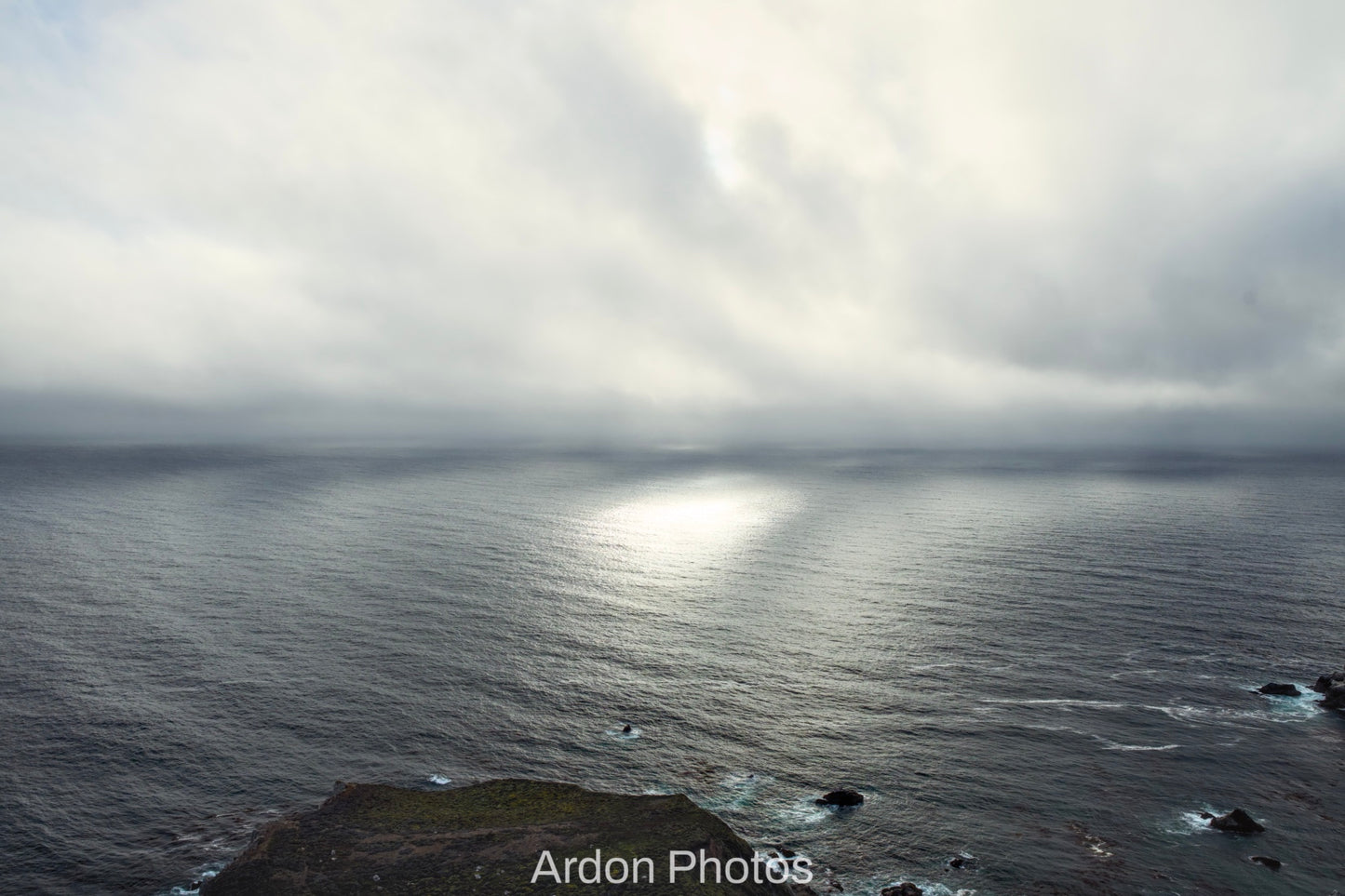 A dramatic coastal seascape with dark ocean water under a cloudy sky, with bright sunlight reflecting on the horizon and a rocky shoreline visible in the foreground. The text 'Ardon Photos' appears at the bottom.