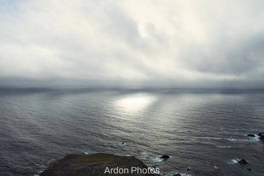 A dramatic coastal seascape with dark ocean water under a cloudy sky, with bright sunlight reflecting on the horizon and a rocky shoreline visible in the foreground. The text 'Ardon Photos' appears at the bottom.
